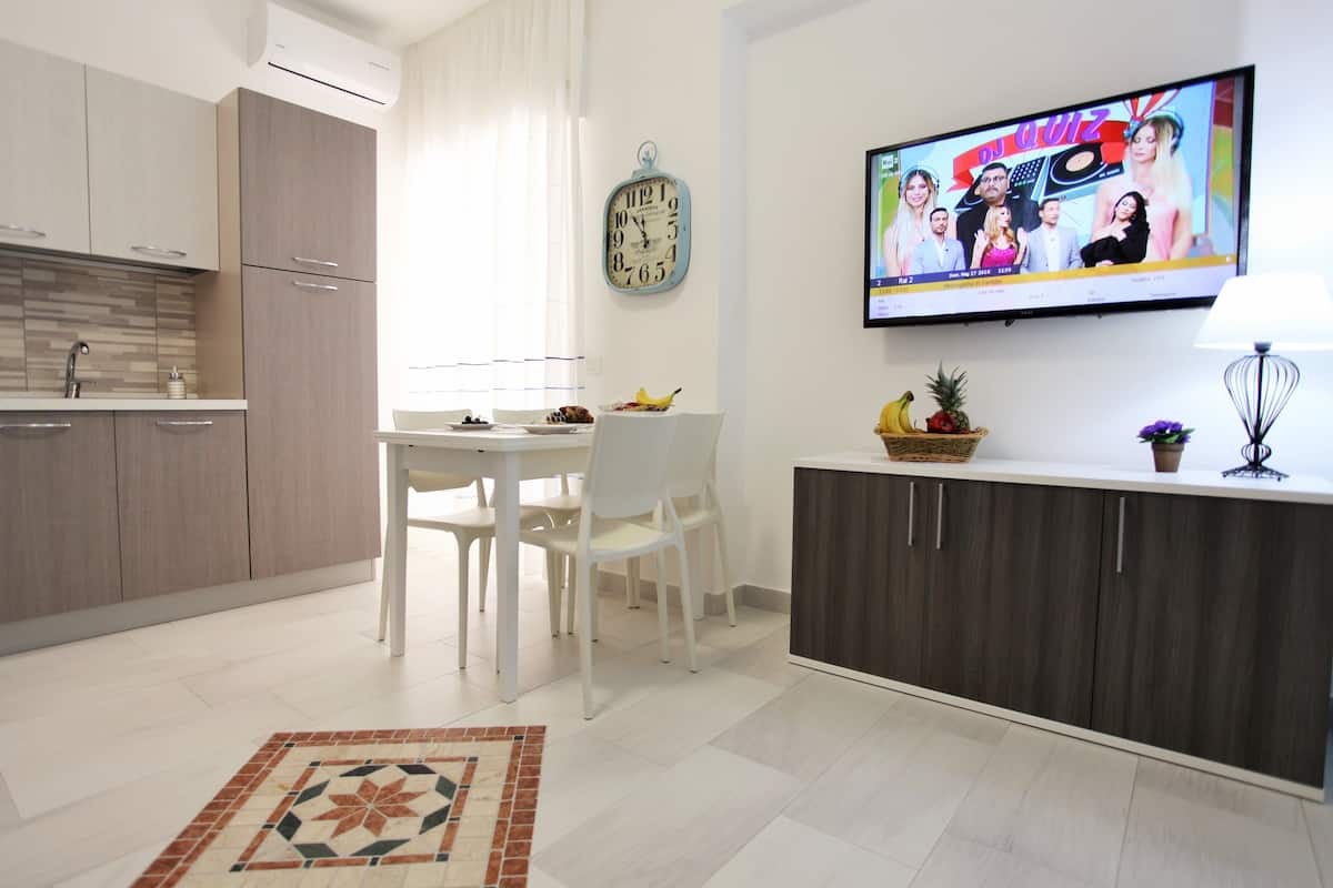A modern kitchen with white cabinets, a dining area with a white table and chairs, a TV mounted on the wall, and a decorative clock.