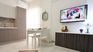 Una cocina moderna con armarios blancos, un comedor con mesa y sillas blancas, un televisor montado en la pared y un reloj decorativo.