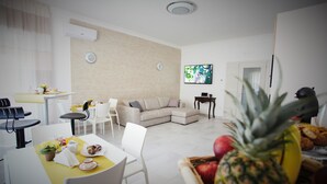 Una sala de estar moderna con sofá, mesa de comedor y sillas. Hay un televisor montado en la pared y una mesita con una computadora portátil.