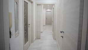 Un pasillo con pisos y paredes de baldosas blancas, puertas con paneles de vidrio e iluminación empotrada en el techo.