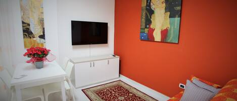 Une pièce avec un bureau blanc, un téléviseur à écran plat, un mur d’accent rouge et un lit avec une literie à motifs.