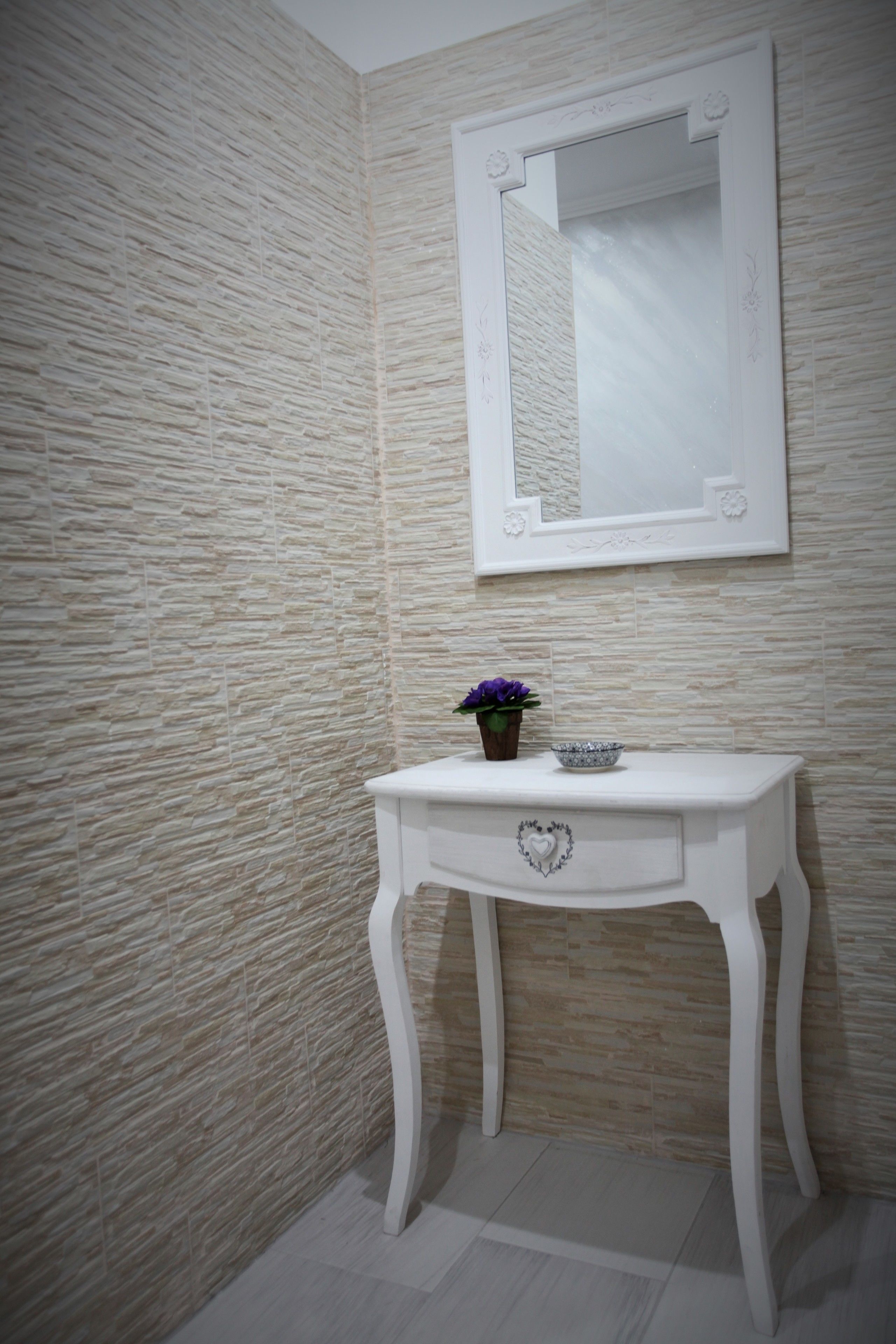 Un baño con pared texturizada, un tocador blanco con cajón en forma de corazón, un espejo y una pequeña planta en maceta.