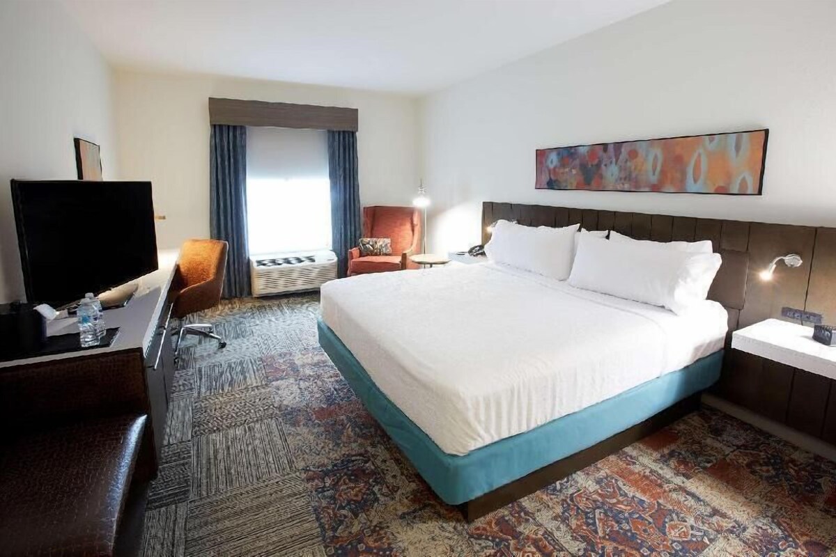 A hotel room with a large bed, a desk, a chair, a TV, and a window with curtains.