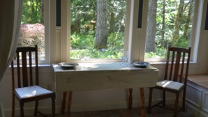 A dining area with a table and two chairs, a window with a view of trees, and a tiled floor. - Two Herons Cottage (Shelton)