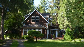A two-story wooden house with a front porch, surrounded by lush greenery and trees. - Two Herons Cottage (Shelton)