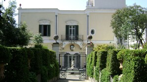 Un edificio con entrada protegida por una reja, una estatua en primer plano y un seto bien cuidado.
