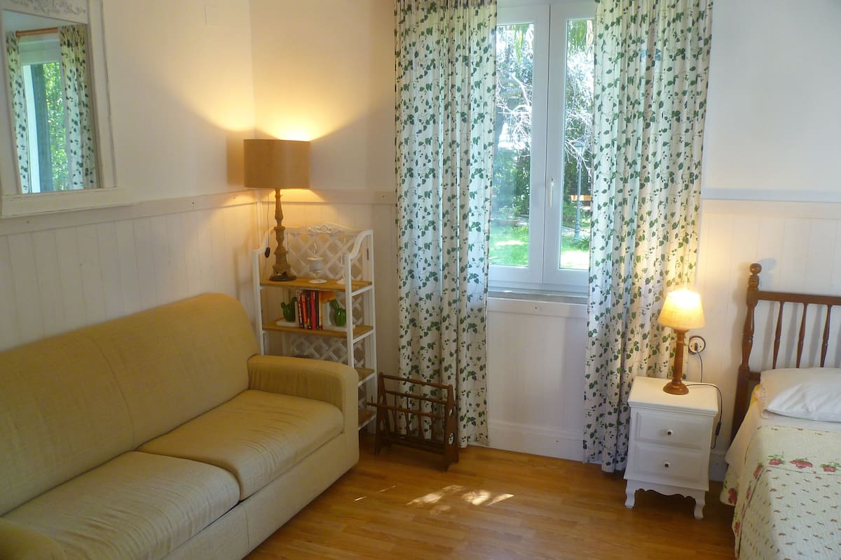Et hyggeligt værelse med trægulv, en beige sofa, et lille hvidt natbord med lampe, en træsenggavl og et vindue med blomstrede gardiner.