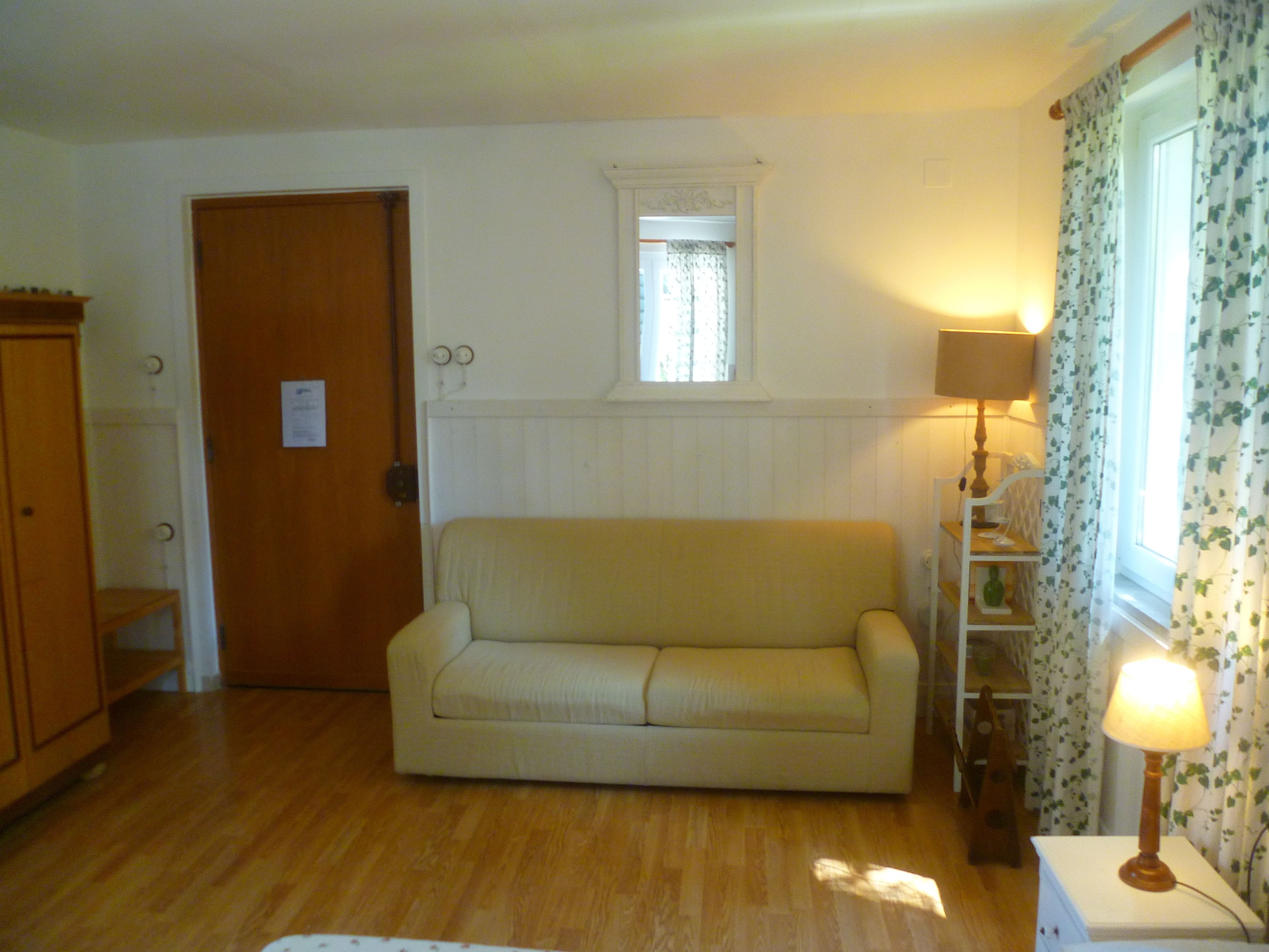 A room with a wooden floor, a beige sofa, a wooden cabinet, a small window with a white frame, and a lamp on a white table.