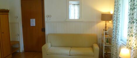 A room with a wooden floor, a beige sofa, a wooden cabinet, a small window with a white frame, and a lamp on a white table.
