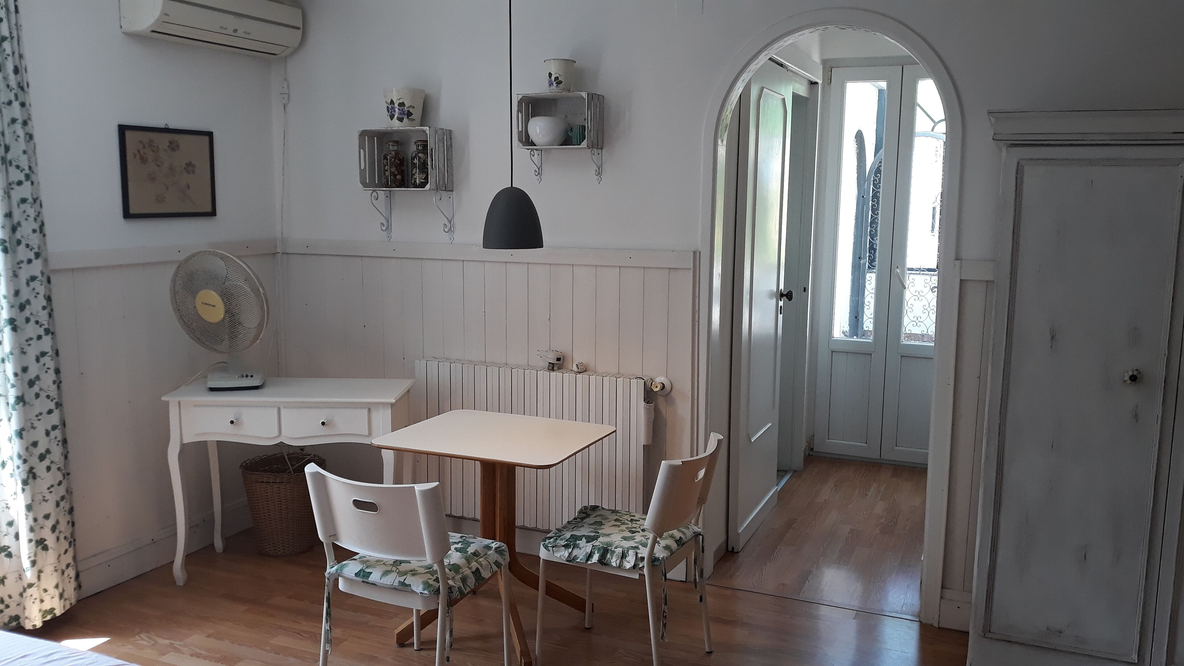 A small dining area with a table and two chairs, a radiator, a wall-mounted shelf with decorative items, and a door leading to another room.