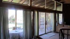 Une pièce dotée de grandes portes-fenêtres coulissantes qui donnent sur un patio extérieur. On y trouve une table ronde recouverte d’une nappe à motifs et une chaise. La pièce est agrémentée d’un plafond en bois avec des poutres apparentes et offre une vue sur l’extérieur grâce aux portes en verre.
