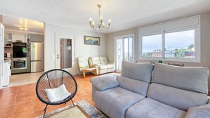 A living room with a grey sofa, a black chair, a chandelier, a dining area with a table and chairs, and a view of the sea through large windows. - RUBENS C1 Almadrava (Rosas / Roses)