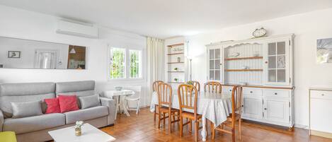 A living room with a grey sofa, a white coffee table, and a dining area with wooden chairs and a white tablecloth.