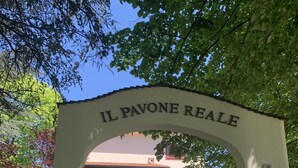 Kaarikuja, jonka yläpuolella on kaiverrus, jossa lukee ”IL PAVONE REALE”, johtaa rakennukseen, jossa on punainen ovi ja valkoinen julkisivu.