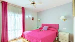 Un dormitorio con cama, mesita de noche, lámpara, ventilador de techo y ventana con cortinas.