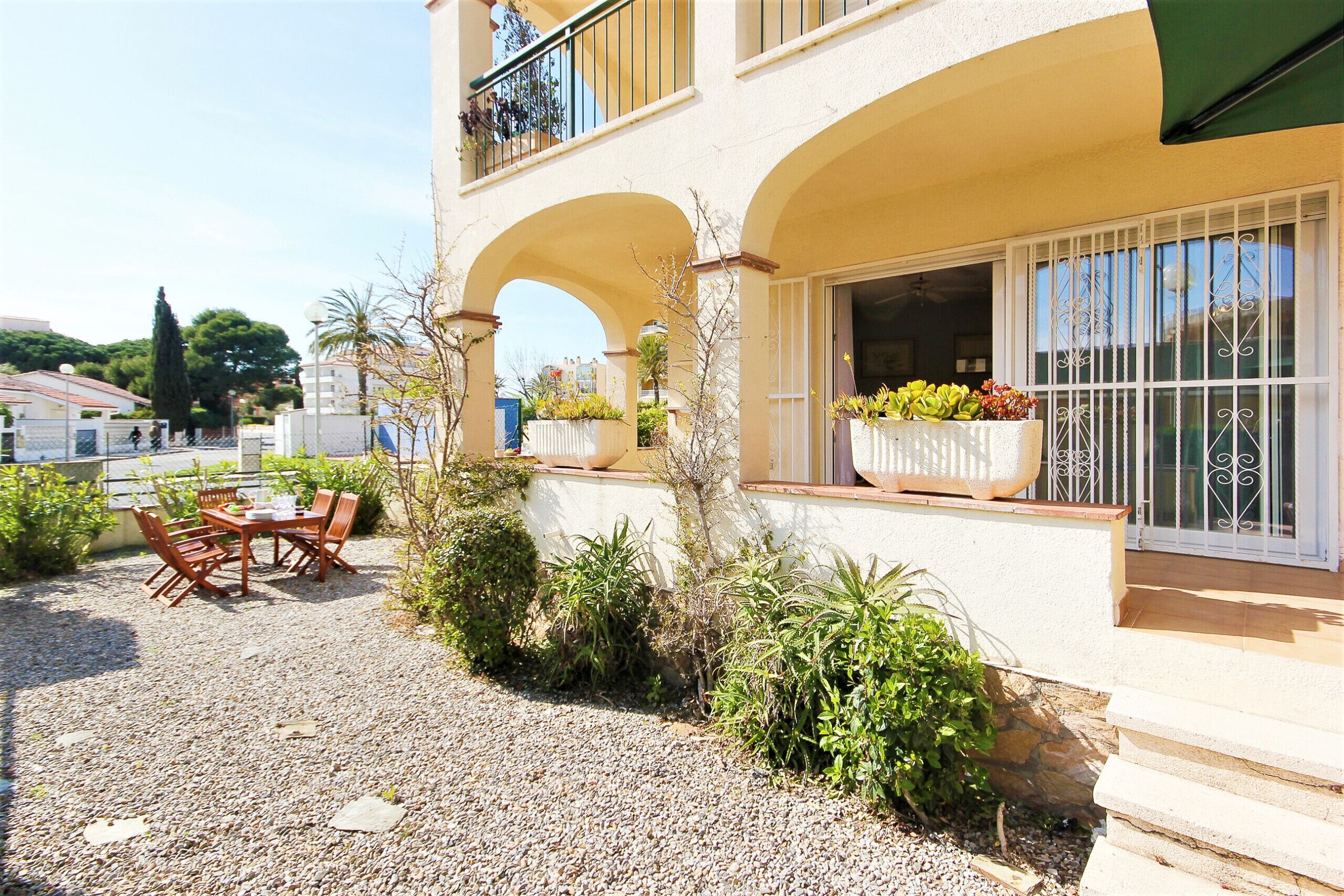 Un edificio de dos pisos con un patio amueblado, con mesa y sillas, rodeado de plantas y un sendero de grava.