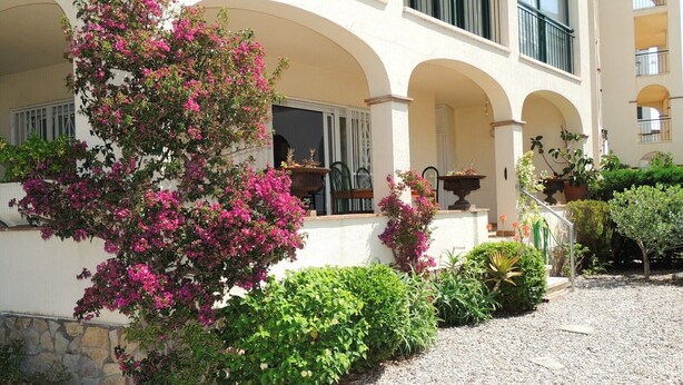 Un edificio con varios balcones, un patio de grava y diversas plantas, incluyendo buganvillas.