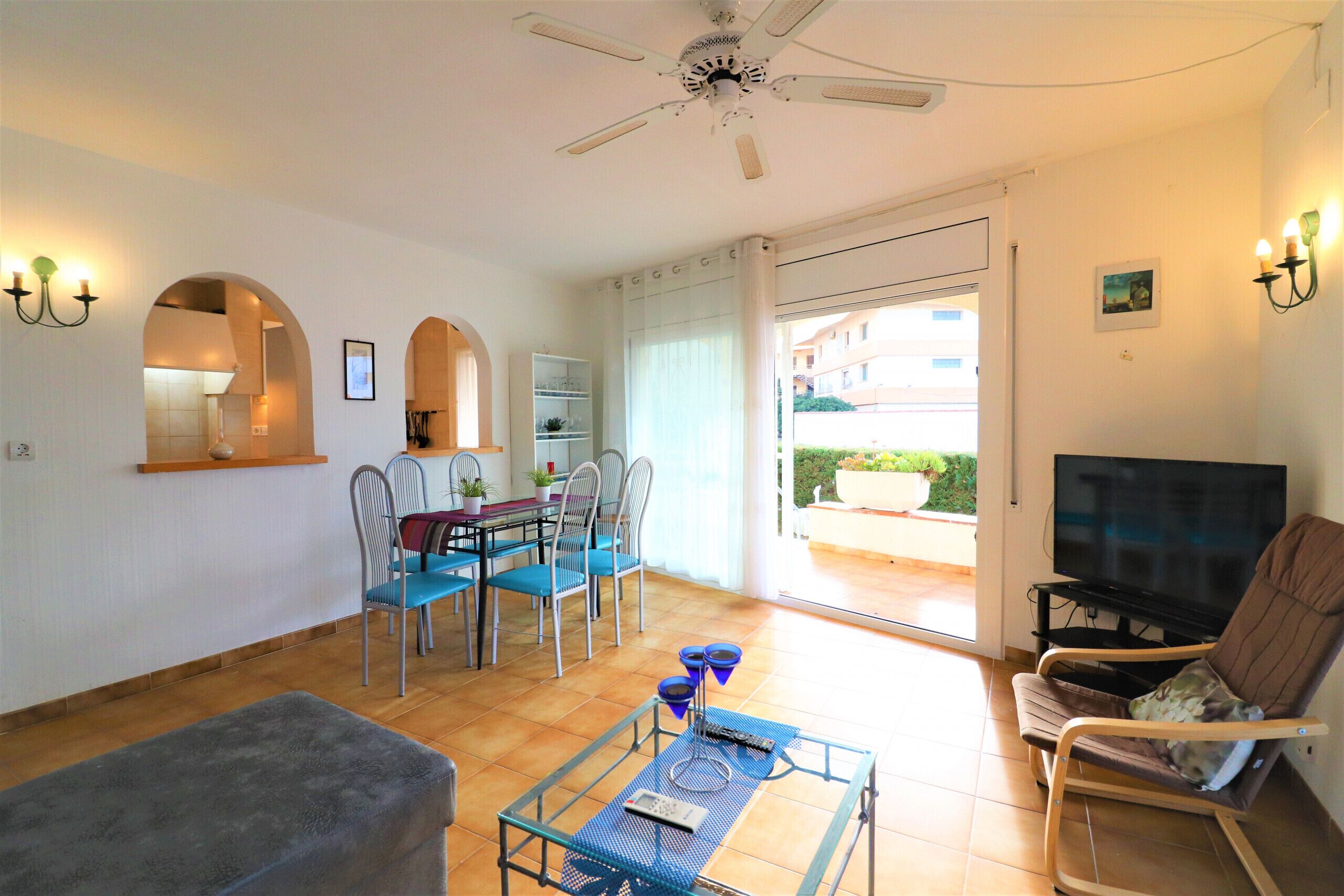 Ein Wohnzimmer mit einer Couch, einem Couchtisch, einem Fernseher und einem Deckenventilator. Es gibt einen Essbereich mit Stühlen und einem Tisch, sowie einen Balkon mit Blick.