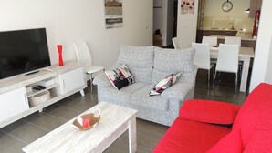 Ett modernt vardagsrum med en platt-TV, en grå soffa med mönstrade kuddar, en röd soffa, ett träbord och en matplats med ett kök synligt i bakgrunden.