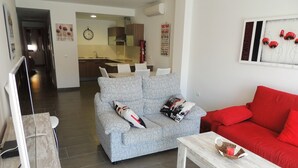 Ett modernt vardagsrum med en grå soffa, en röd soffa och ett soffbord i trä. En platt-TV står på ett TV-bord, och i bakgrunden syns en köksdel samt en väggklocka.