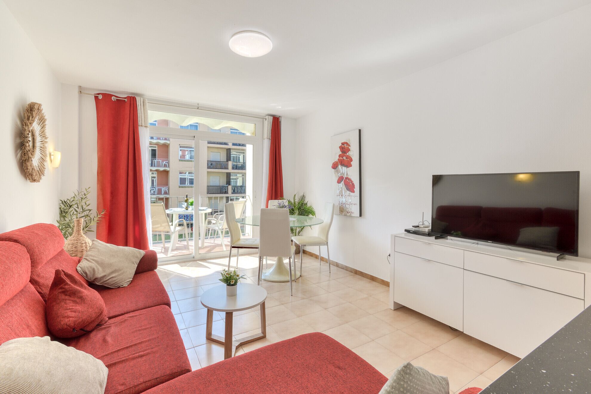 A living room with a sofa, a TV, a dining area with chairs and a table, and a balcony with a view of buildings.