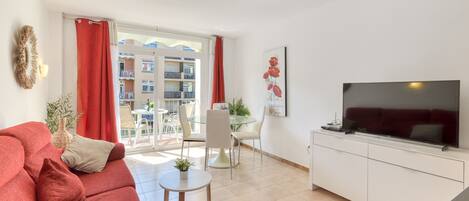 A living room with a sofa, a TV, a dining area with chairs and a table, and a balcony with a view of buildings.