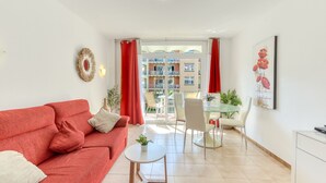 A living room with a red sofa, a small round table, and a glass dining table with chairs. There is a balcony with red curtains and a view of buildings.