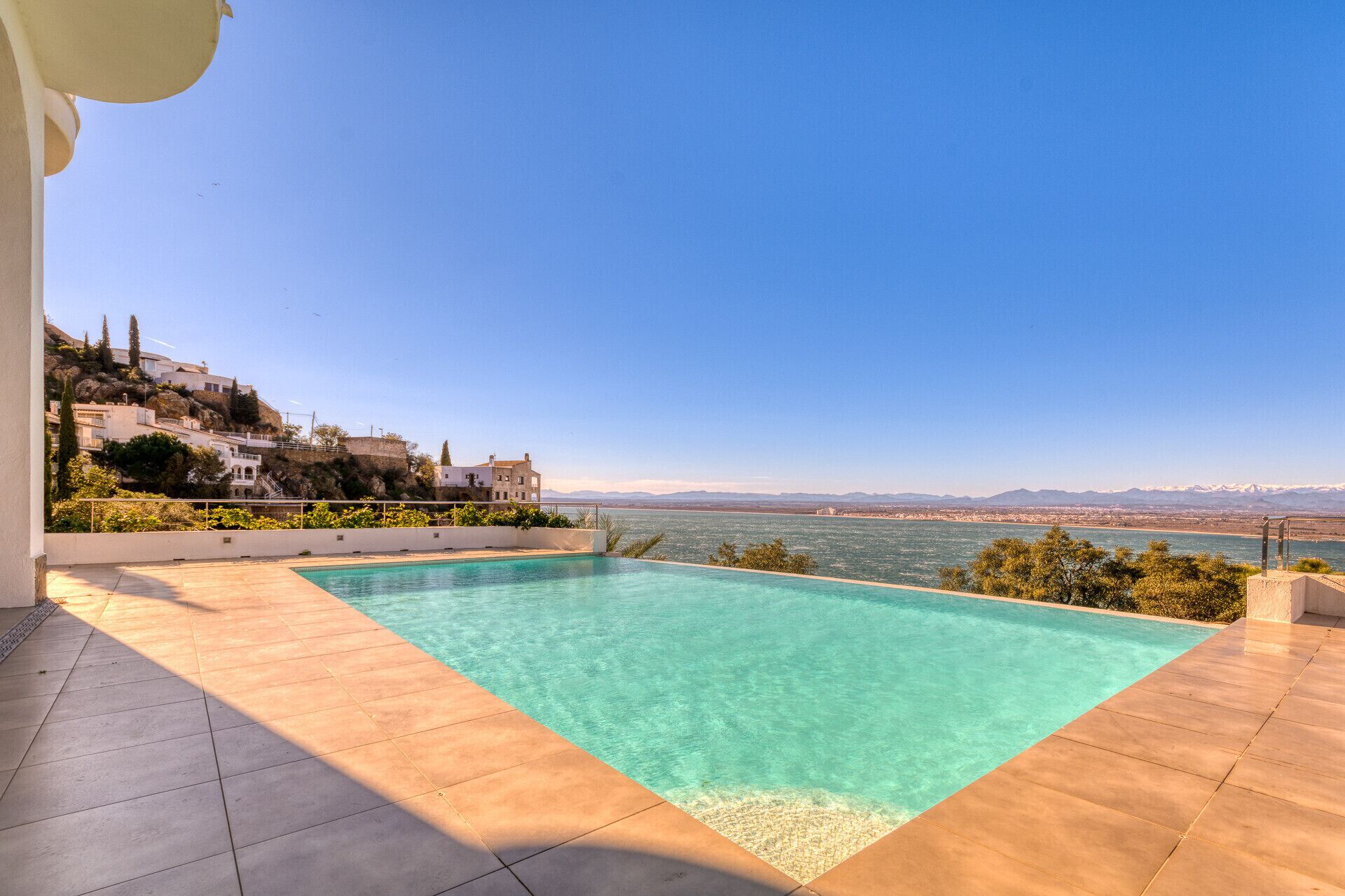 A swimming pool with clear water, a tiled deck, and a view of a coastal landscape with buildings.