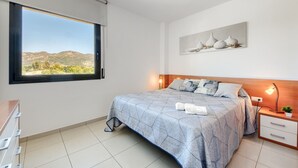 A bedroom with a large window, a bed with a patterned bedspread, a bedside table with a lamp, and a wall-mounted artwork.
