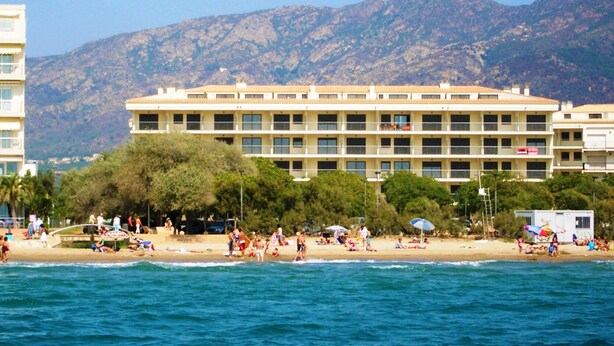 A coastal hotel with multiple floors, a beach in the foreground, and mountains in the background.