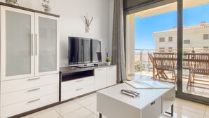 A modern living room with a flat-screen TV, a small table with a remote, and a balcony with wooden furniture.
