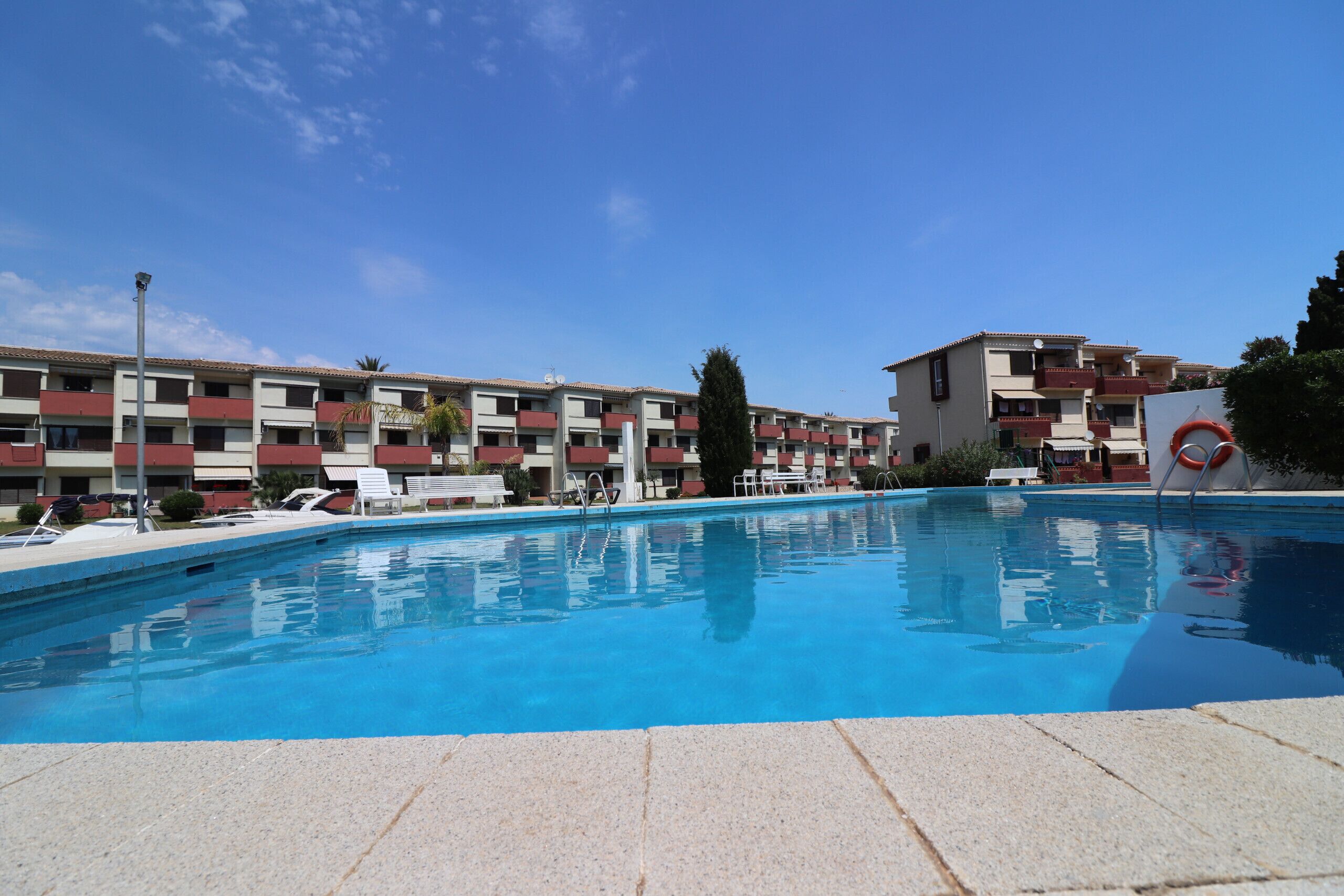 Una piscina con lettini, una postazione di salvataggio e un edificio con balconi sullo sfondo.