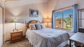 Une chambre à coucher avec un lit, une table de chevet, une lampe, une fenêtre donnant sur l’extérieur et un tableau accroché au mur.