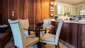 Een eethoek met een glazen tafel en vier stoelen, een houten wand en een keuken op de achtergrond.