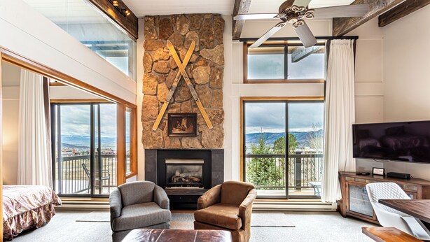 A spacious living room with a stone fireplace, large windows, and a balcony with a view.