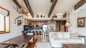 A modern kitchen with wooden cabinets, a dining area with a wooden table and benches, a white sofa with patterned cushions, and a rustic chandelier.