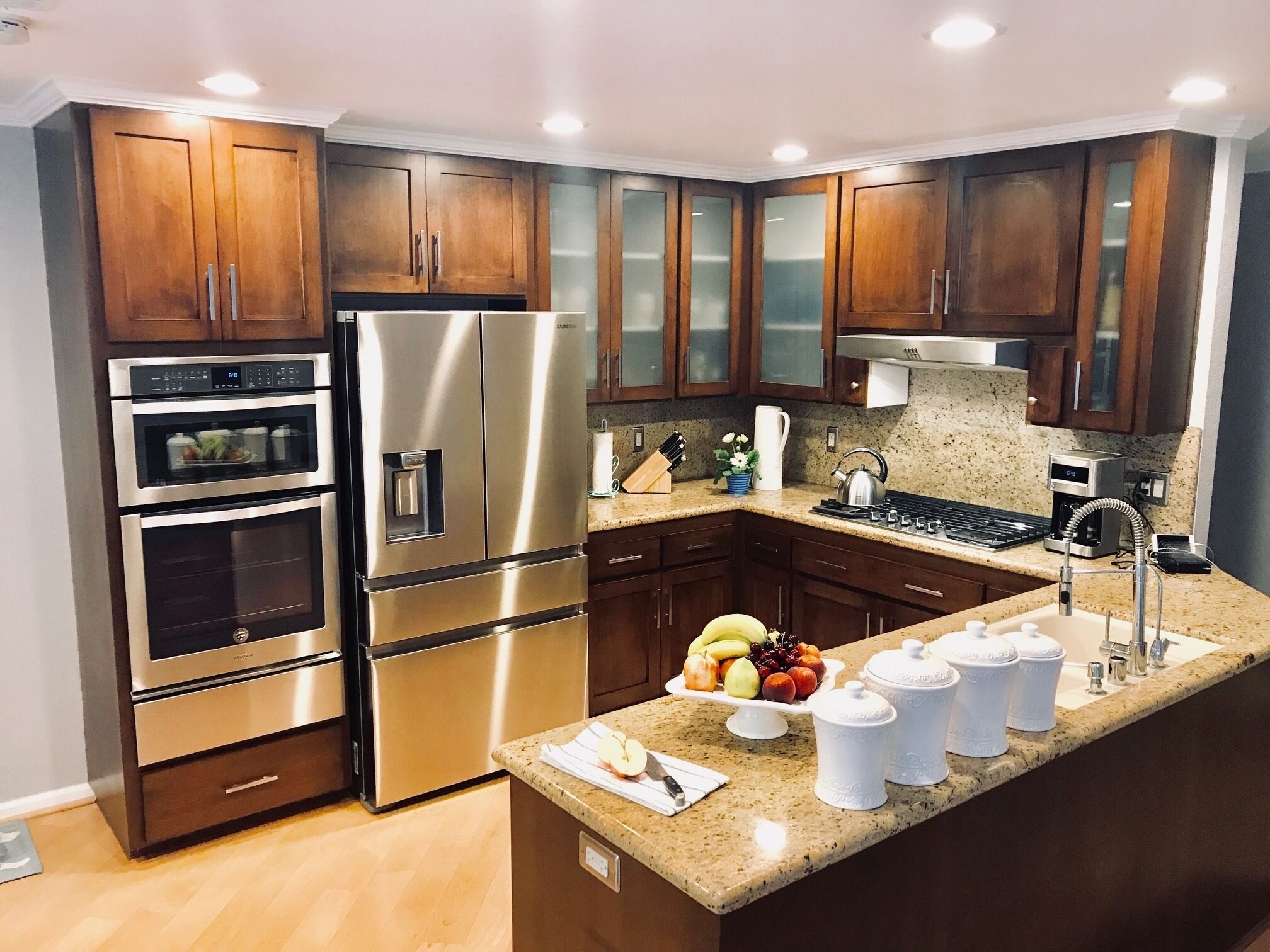 A modern kitchen with stainless steel appliances, wooden cabinets, granite countertops, and a central island with fruit and storage containers.