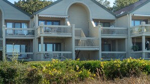 A two-story building with balconies, a central archway, and a garden area with yellow flowers.