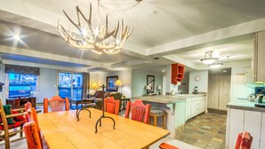 Uma sala de estar ampla com uma mesa de jantar de madeira, cadeiras vermelhas, um candelabro e uma cozinha ao fundo.