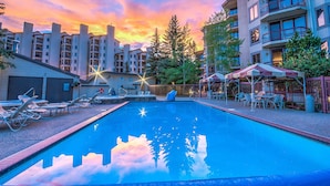 Piscina com espreguiçadeiras e guarda-sóis, rodeada por edifícios altos e árvores.