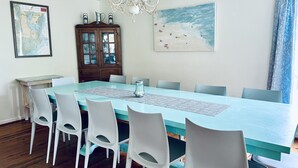 Uma sala de jantar com uma mesa azul clara, cadeiras brancas, um armário de madeira, um candelabro, um mapa e uma pintura de praia.