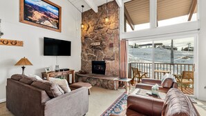 Un salon chaleureux avec une cheminée en pierre, des fauteuils confortables, un téléviseur à écran plat et une vue sur un paysage enneigé grâce à de grandes fenêtres.