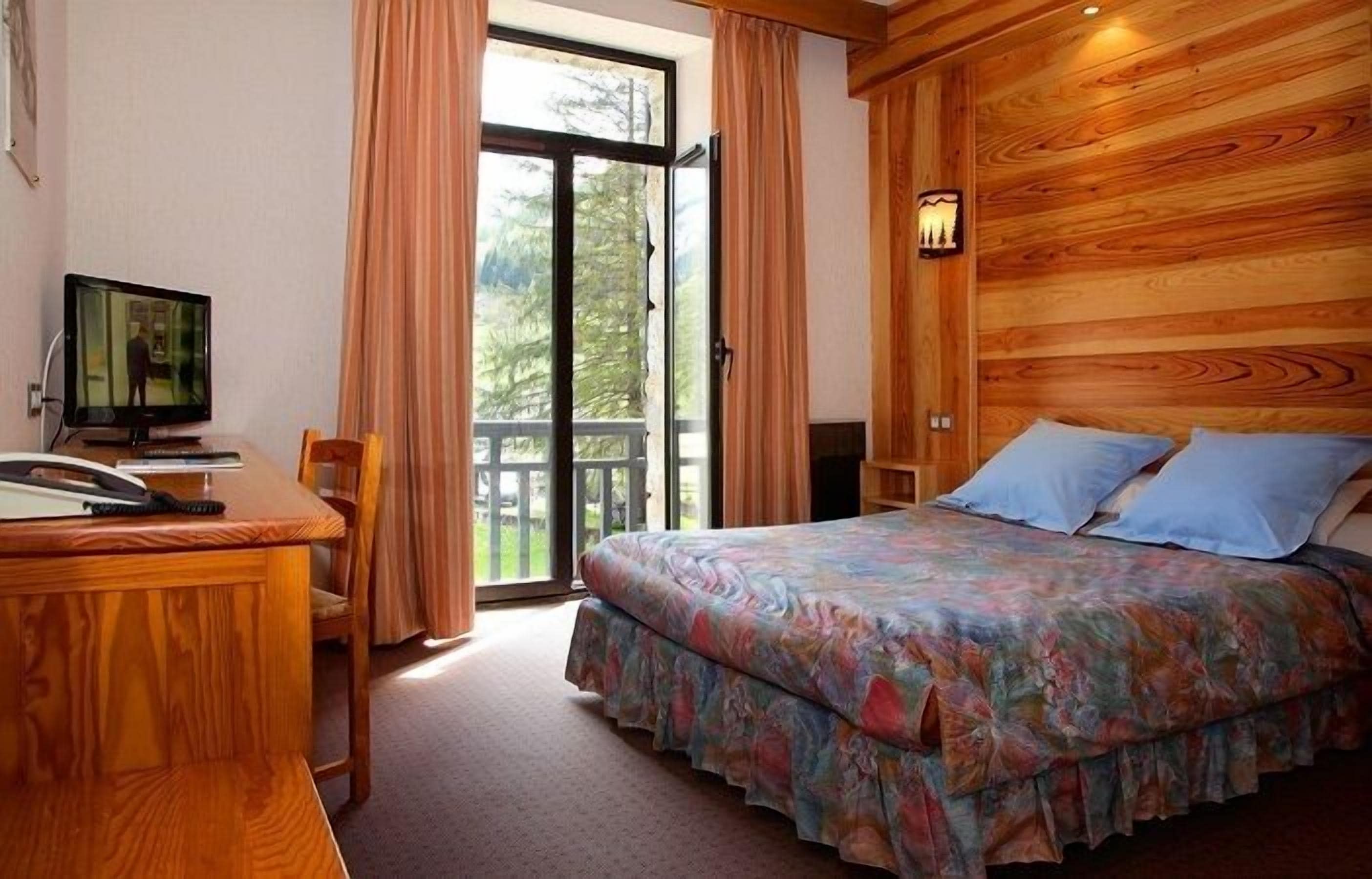 A bedroom with a bed, a desk, a television, and a window with curtains.