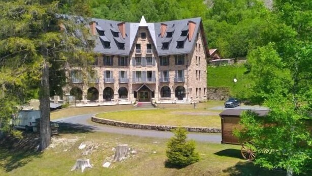 A large stone building with multiple balconies, surrounded by lush greenery and a river. - Hôtel Vignemale (Gavarnie-Gedre)