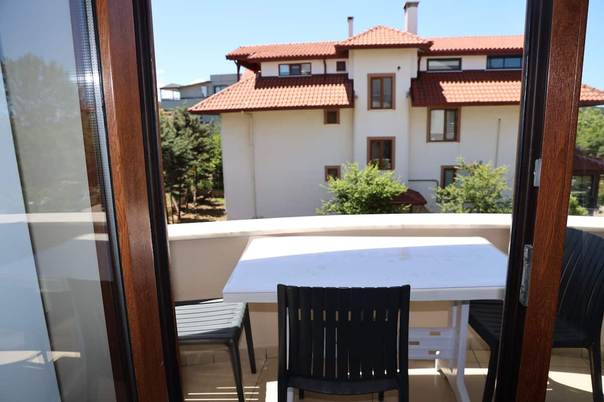 A balcony with a table and chairs, a view of a house with a red-tiled roof, and a clear sky.
