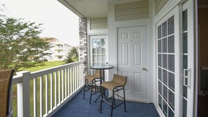 A balcony with two chairs and a table, a white door, and a view of a building across the lawn. - 4673 Wild Iris Dr 203 2 Bedroom Condo by Redawning (Myrtle Beach)