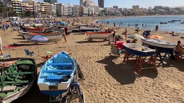 A beach with multiple boats and umbrellas, a sandy shore, and buildings in the background. - Casa a Medida (Las Palmas de Gran Canaria)