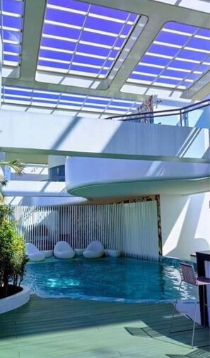 Un espace piscine intérieur moderne doté d’un toit en verre, de plusieurs niveaux et d’un coin salon avec des sièges.