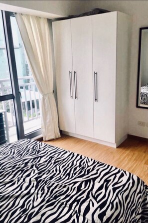 Une chambre à coucher avec une couvre-lit à motifs zébrés, une armoire blanche à deux portes et une grande fenêtre avec un rideau.