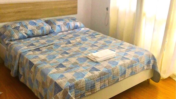 Una cama bien hecha con colcha y almohadas a cuadros azules y blancos, un cabecero de madera y una ventana con cortinas.